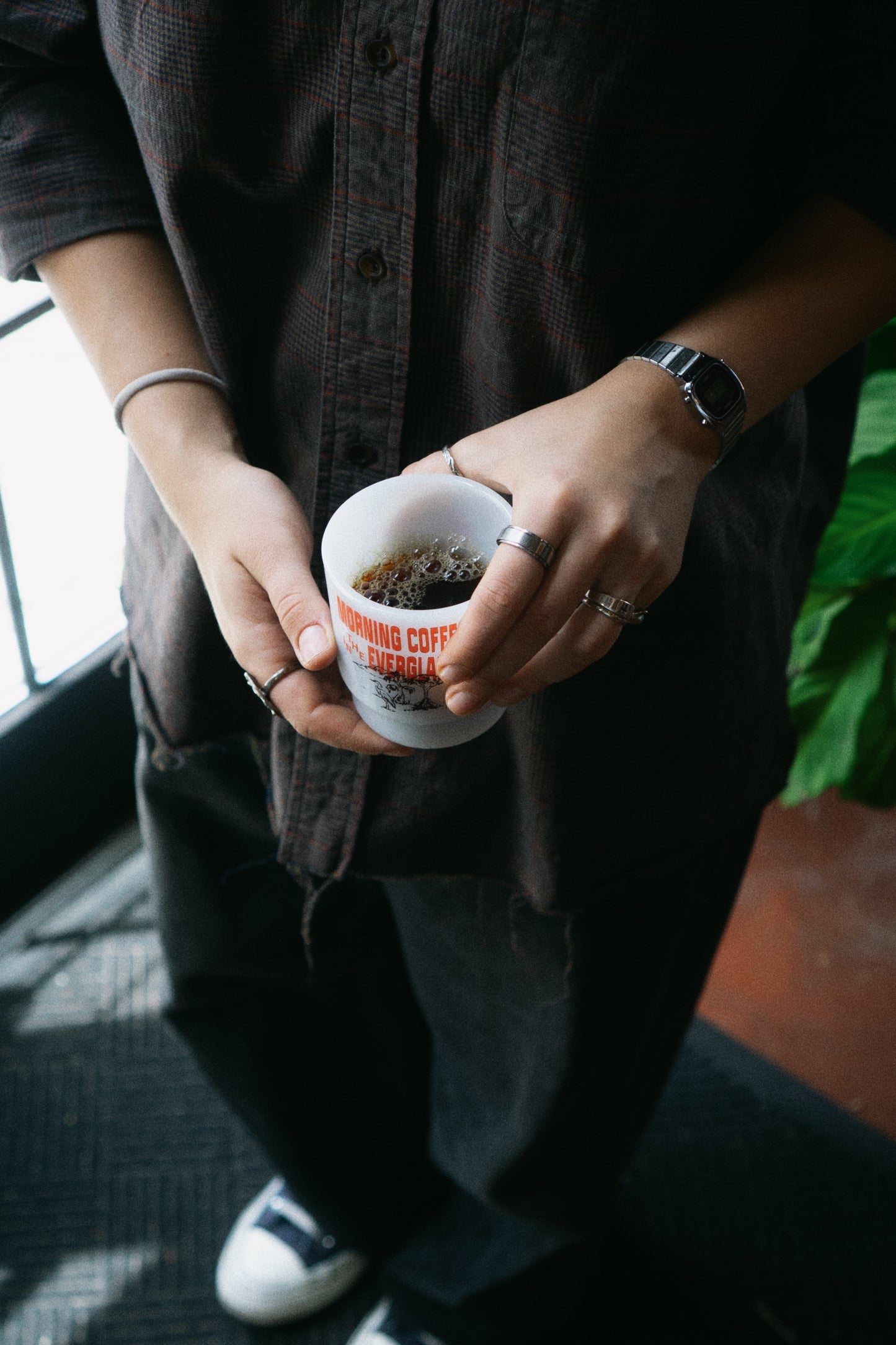 A person wearing a dark brown plaid button-up shirt and black pants holds a white ceramic coffee cup printed with 'MORNING COFFEE IN THE EVERGLADES' and red text, standing indoors near a window.