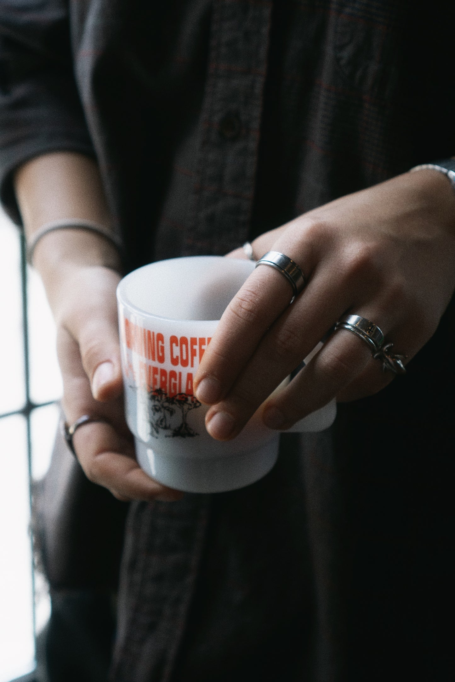 A person in a dark button-up shirt holds a white ceramic coffee mug with orange text that reads 'MAKING COFFEE IN THE EVERGLADES'. The person is wearing multiple silver rings on their fingers.