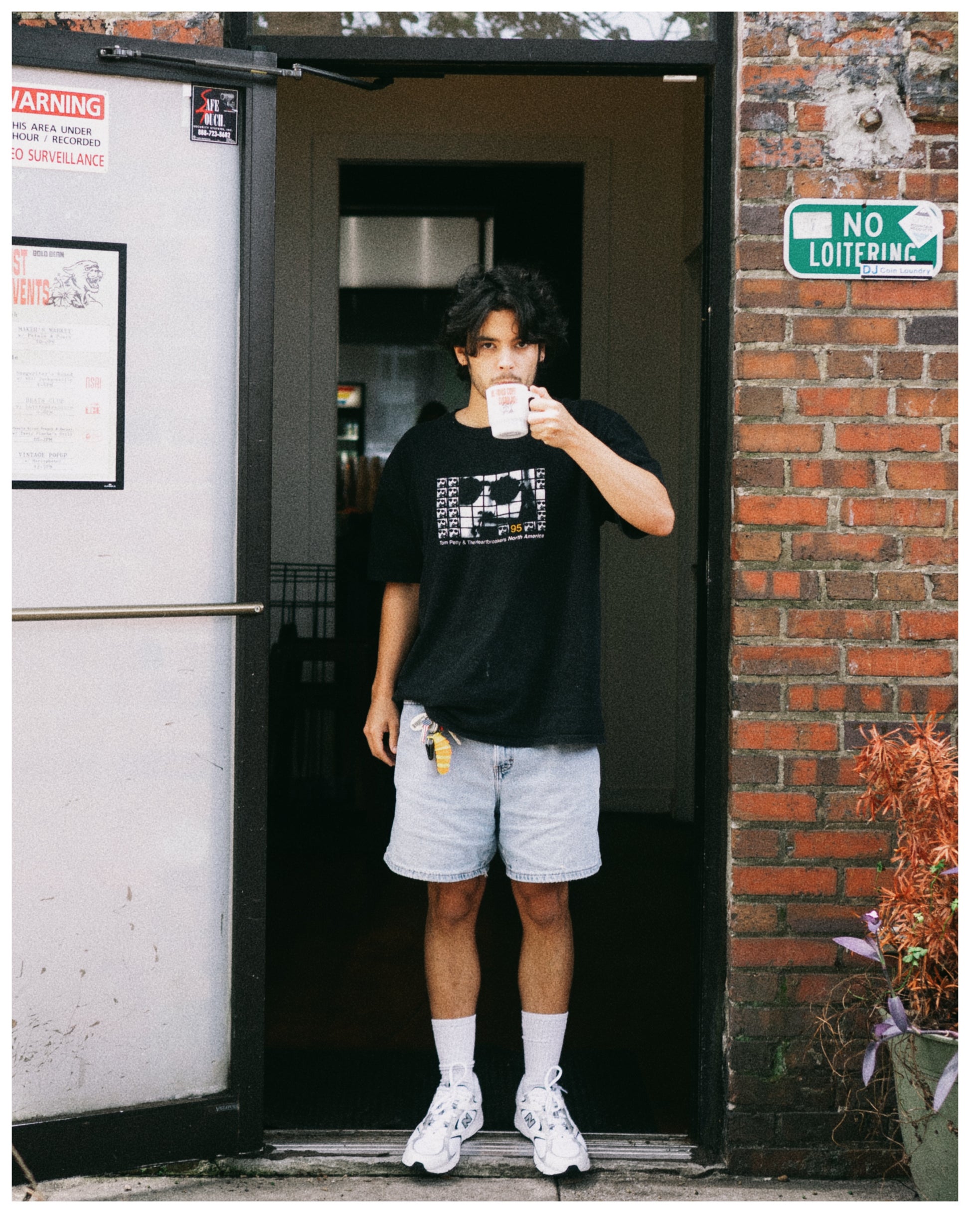 A man stands in a doorway wearing a black graphic t-shirt, light gray shorts, white socks, and white sneakers, holding a white mug. The background features a brick wall on one side and a white door on the other.