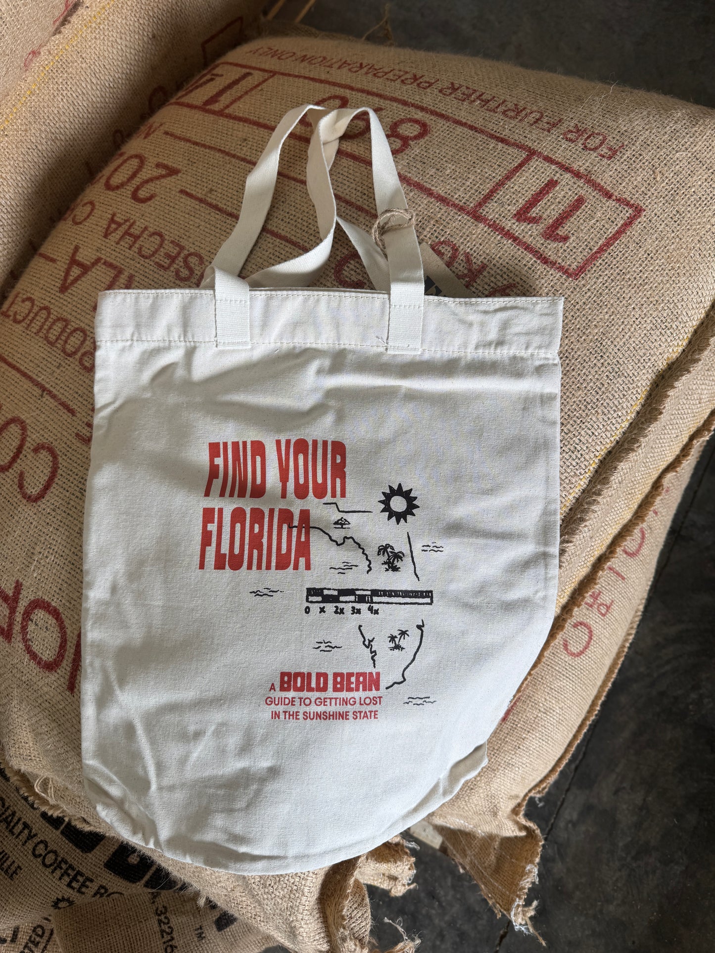 A white canvas tote bag with red and black graphic text reading 'FIND YOUR FLORIDA' and 'A BOLD BERN' lies on top of large brown burlap sacks.