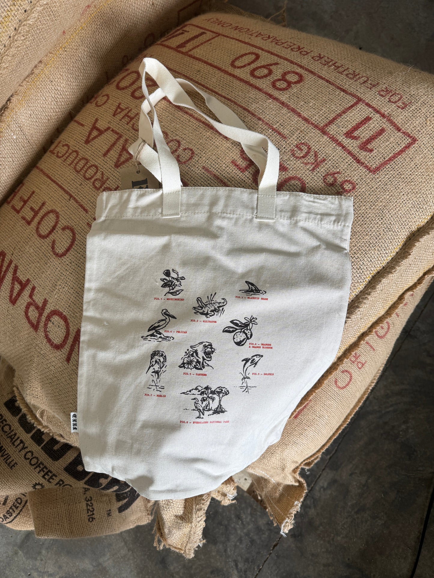 A white canvas tote bag with printed illustrations and small red labels, resting on top of stacked burlap sacks printed with red coffee text.