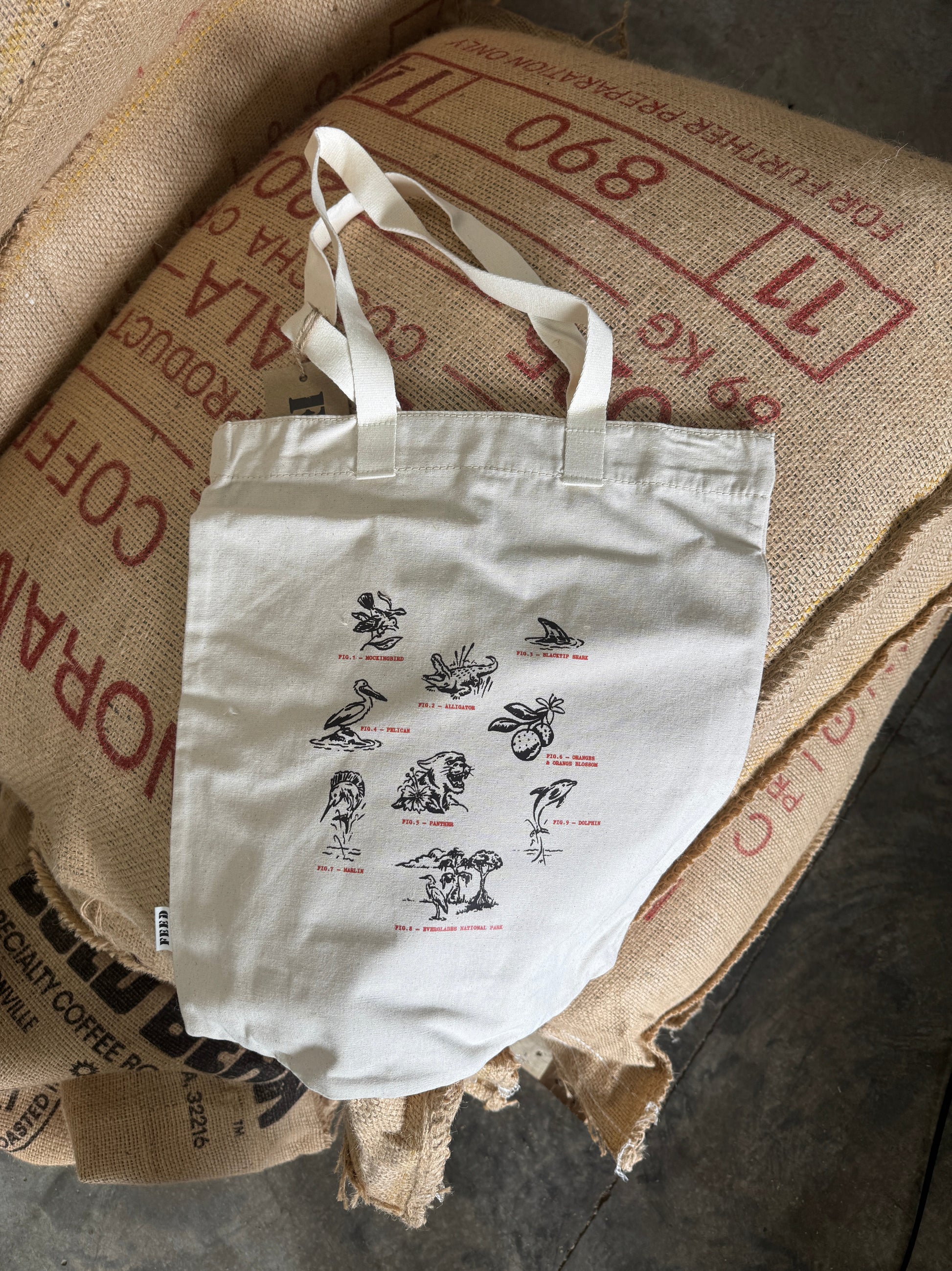 A white canvas tote bag with printed illustrations and small red labels, resting on top of stacked burlap sacks printed with red coffee text.