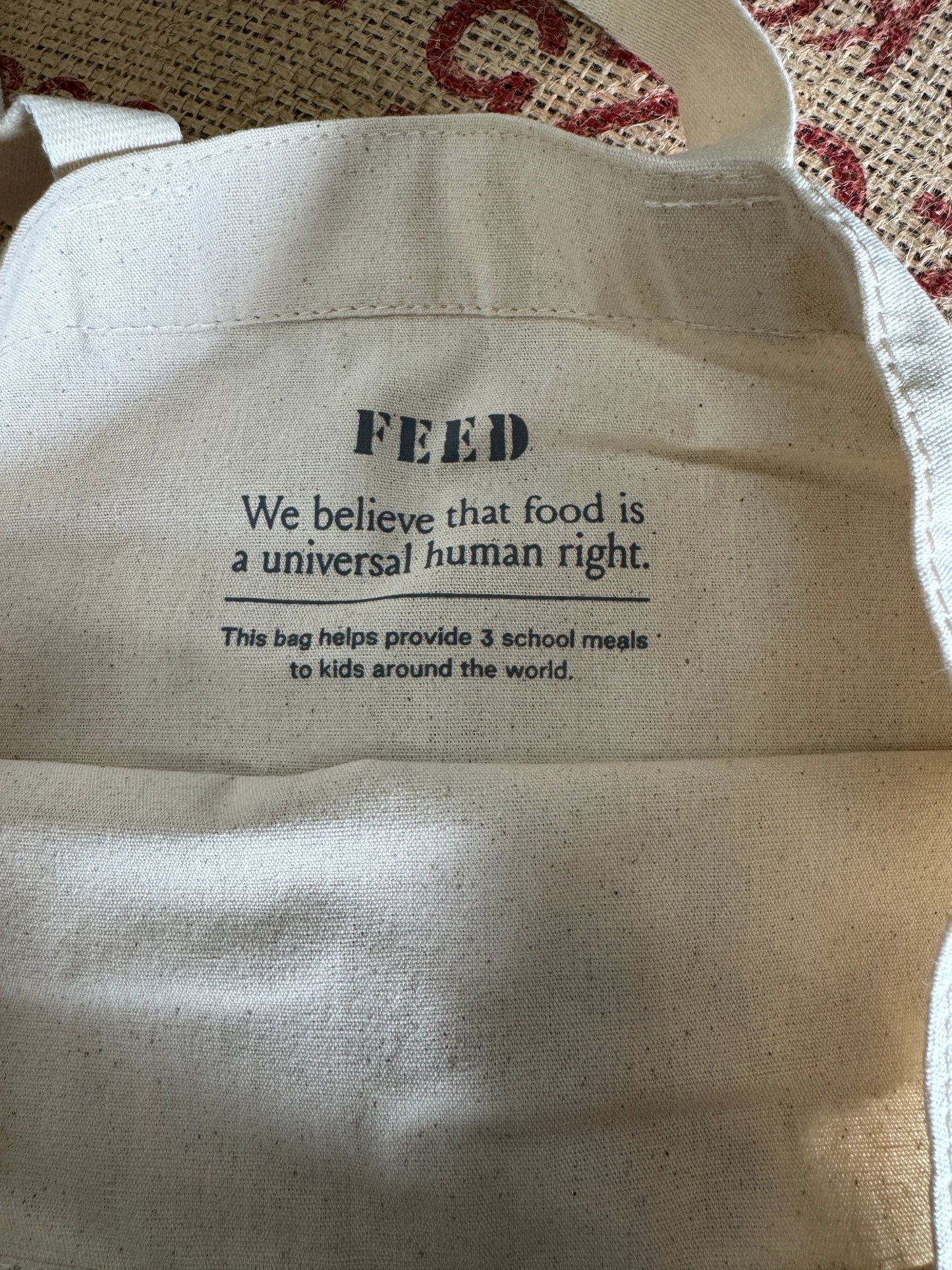 A close-up of a beige canvas tote bag with black printed text on the inside. The text reads 'FEED We believe that food is a universal human right.