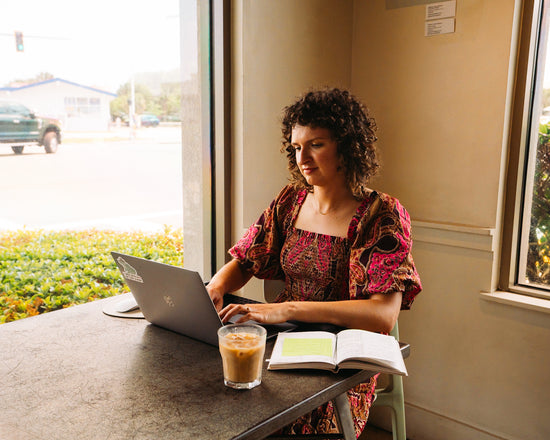 image of curly haired woman in decorative dress sitting at a table in a coffee shop working on computer.  A glass of iced coffee is on the table along with a book