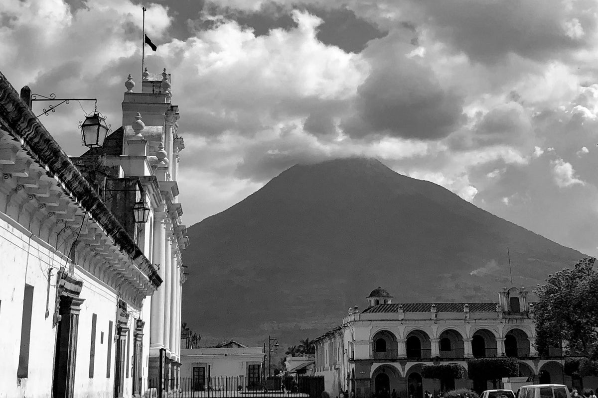 Black and white photo of a city street with cars and buildings under a cloudy sky with volcano in background.